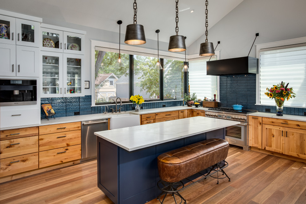 This is an example of a large rural l-shaped open plan kitchen in Denver with a belfast sink, shaker cabinets, quartz worktops, blue splashback, glass tiled splashback, stainless steel appliances, medium hardwood flooring, an island, white worktops, a vaulted ceiling, medium wood cabinets and brown floors.