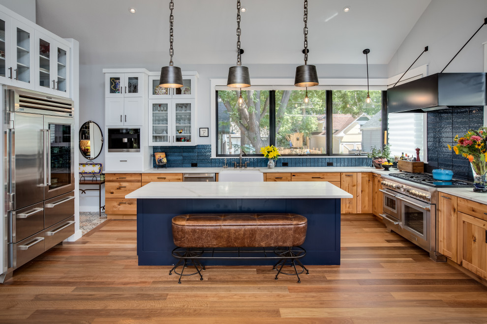 This is an example of a large farmhouse l-shaped open plan kitchen in Denver with a belfast sink, shaker cabinets, light wood cabinets, quartz worktops, blue splashback, glass tiled splashback, stainless steel appliances, medium hardwood flooring, an island, white worktops and a vaulted ceiling.