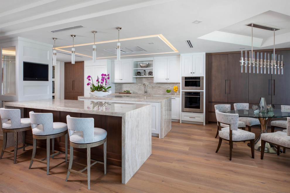 Example of a large beach style medium tone wood floor and brown floor eat-in kitchen design in Miami with an undermount sink, recessed-panel cabinets, white cabinets, quartzite countertops, beige backsplash, stone slab backsplash, stainless steel appliances, two islands and beige countertops