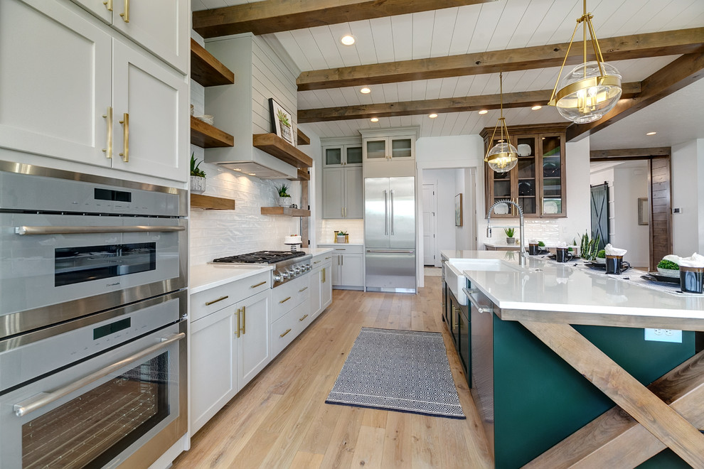 Photo of a large farmhouse l-shaped open plan kitchen in Boise with a belfast sink, shaker cabinets, white cabinets, engineered stone countertops, white splashback, metro tiled splashback, stainless steel appliances, light hardwood flooring, an island, beige floors and white worktops.