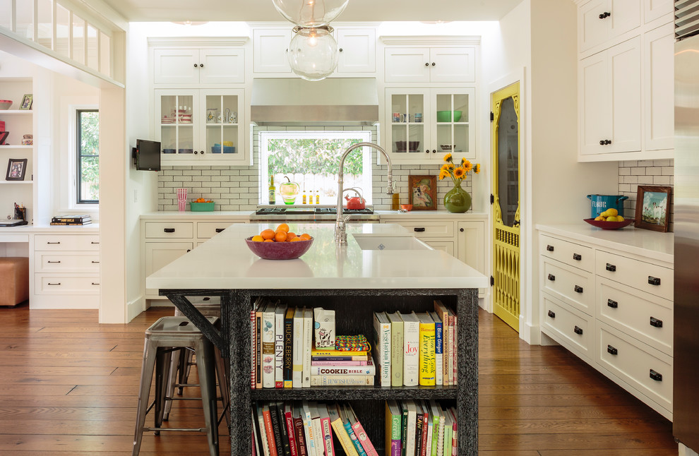 Large farmhouse open plan kitchen in Los Angeles with a belfast sink, shaker cabinets, engineered stone countertops, white splashback, brick splashback, medium hardwood flooring, an island, stainless steel appliances and white cabinets.