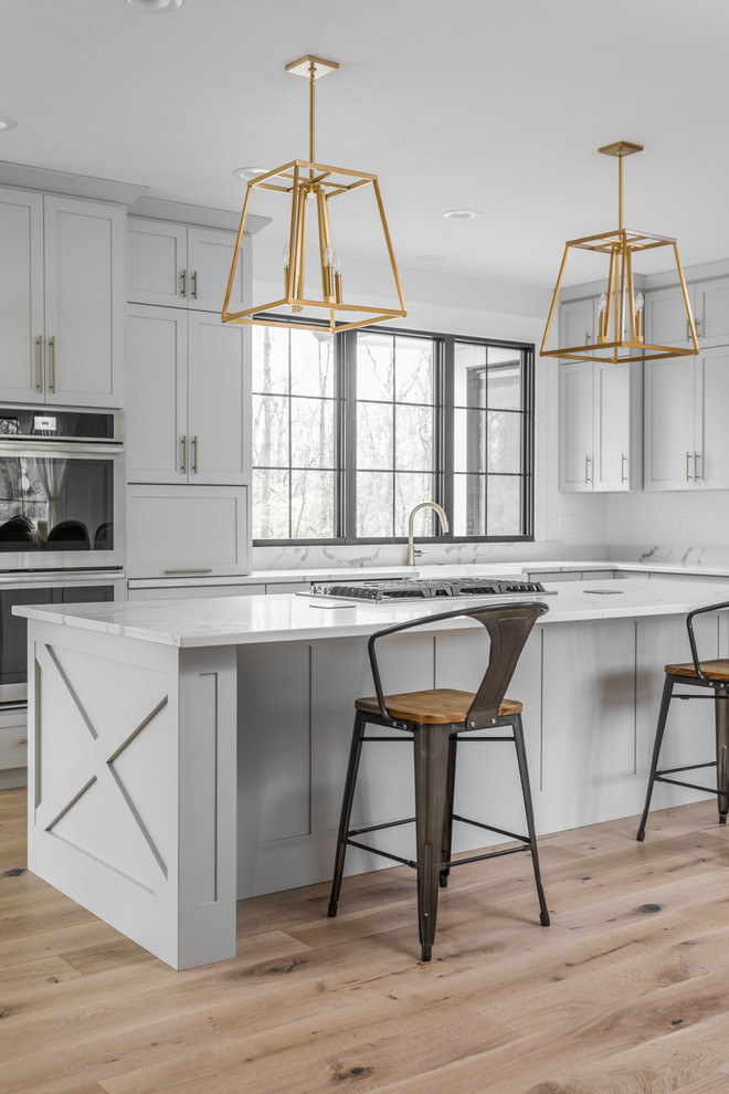 Design ideas for a farmhouse l-shaped kitchen in Indianapolis with shaker cabinets, grey cabinets, white splashback, stainless steel appliances, light hardwood flooring, an island, beige floors and white worktops.