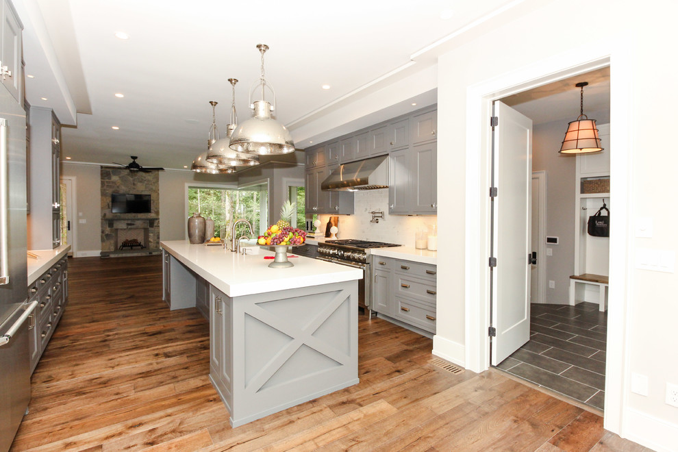 Foto de cocinas en U campestre de tamaño medio cerrado con fregadero sobremueble, armarios estilo shaker, puertas de armario grises, encimera de acrílico, salpicadero blanco, electrodomésticos de acero inoxidable, suelo de madera en tonos medios y una isla