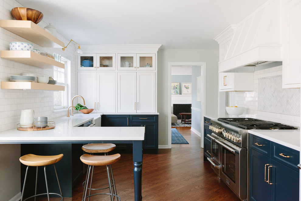 Farmhouse u-shaped kitchen in Indianapolis with a breakfast bar, a belfast sink, shaker cabinets, blue cabinets, white splashback, brown floors, white worktops, stainless steel appliances and medium hardwood flooring.