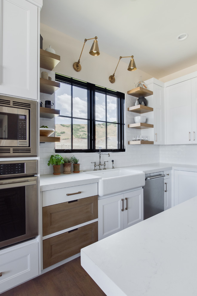 This is an example of a farmhouse l-shaped kitchen in Salt Lake City with a belfast sink, white cabinets, white splashback, metro tiled splashback, stainless steel appliances, light hardwood flooring, an island, white worktops, quartz worktops and recessed-panel cabinets.