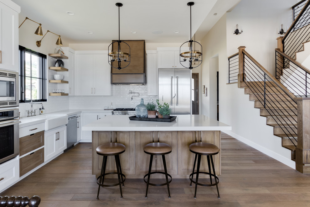 Photo of a country l-shaped kitchen in Salt Lake City with a belfast sink, shaker cabinets, white cabinets, white splashback, metro tiled splashback, stainless steel appliances, light hardwood flooring, an island, white worktops and quartz worktops.