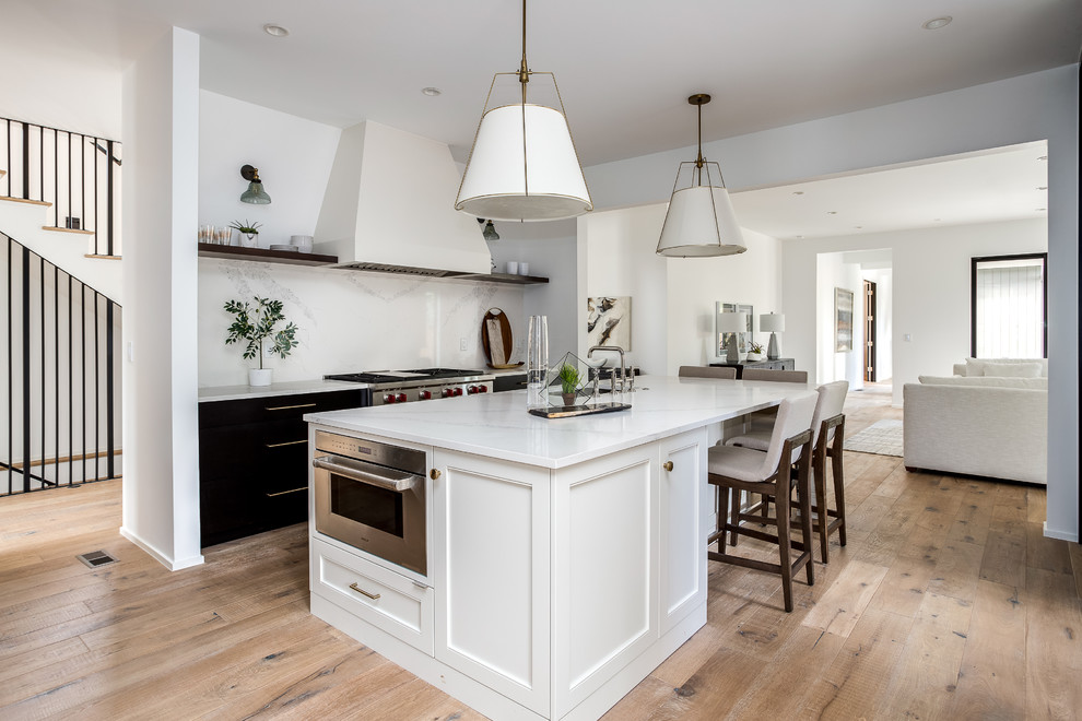 This is an example of a large rural galley kitchen in Denver with a belfast sink, shaker cabinets, white cabinets, marble worktops, white splashback, marble splashback, stainless steel appliances, light hardwood flooring, an island, brown floors and white worktops.
