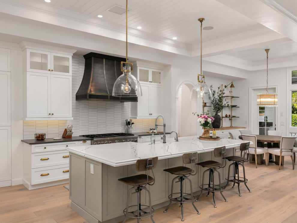 Photo of a country galley kitchen in Orlando with a belfast sink, shaker cabinets, white cabinets, white splashback, stainless steel appliances, medium hardwood flooring, an island, brown floors and white worktops.