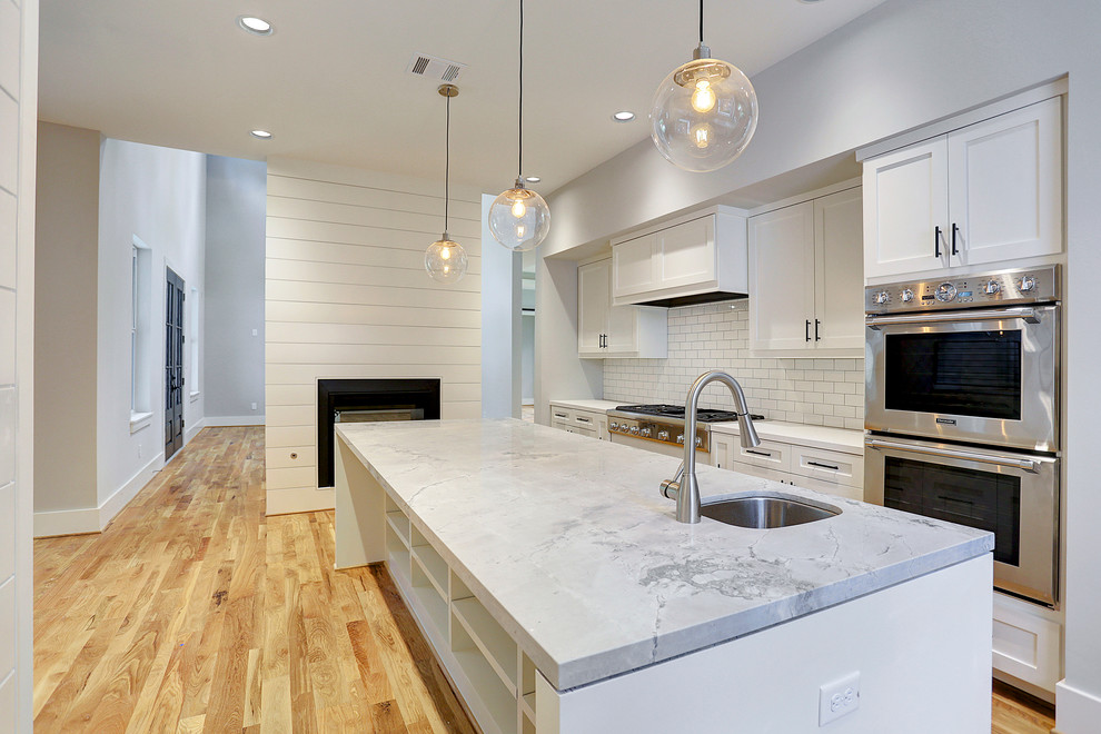 Photo of a large contemporary u-shaped open plan kitchen in Houston with a belfast sink, recessed-panel cabinets, white cabinets, marble worktops, white splashback, porcelain splashback, stainless steel appliances, light hardwood flooring, an island and beige floors.