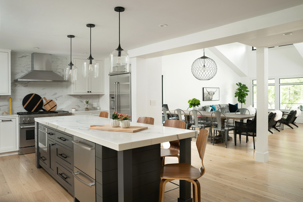 Bild på ett lantligt kök, med en rustik diskho, skåp i shakerstil, vita skåp, bänkskiva i kvarts, stänkskydd i porslinskakel, rostfria vitvaror, ljust trägolv och en köksö