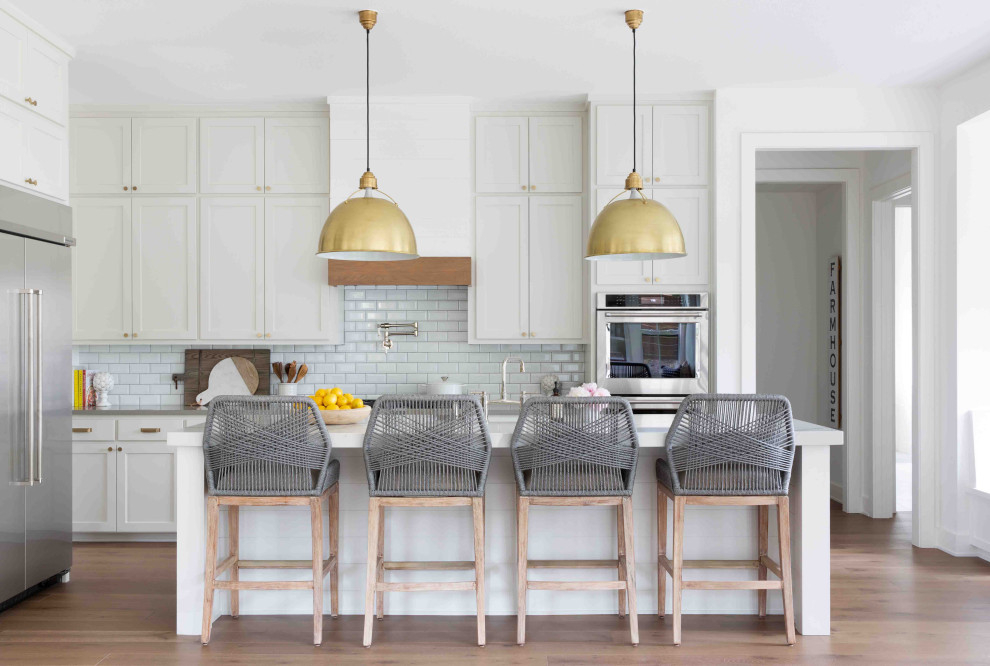 Inspiration for a large country l-shaped kitchen in Austin with shaker cabinets, white cabinets, grey splashback, metro tiled splashback, stainless steel appliances, medium hardwood flooring, an island, brown floors and white worktops.