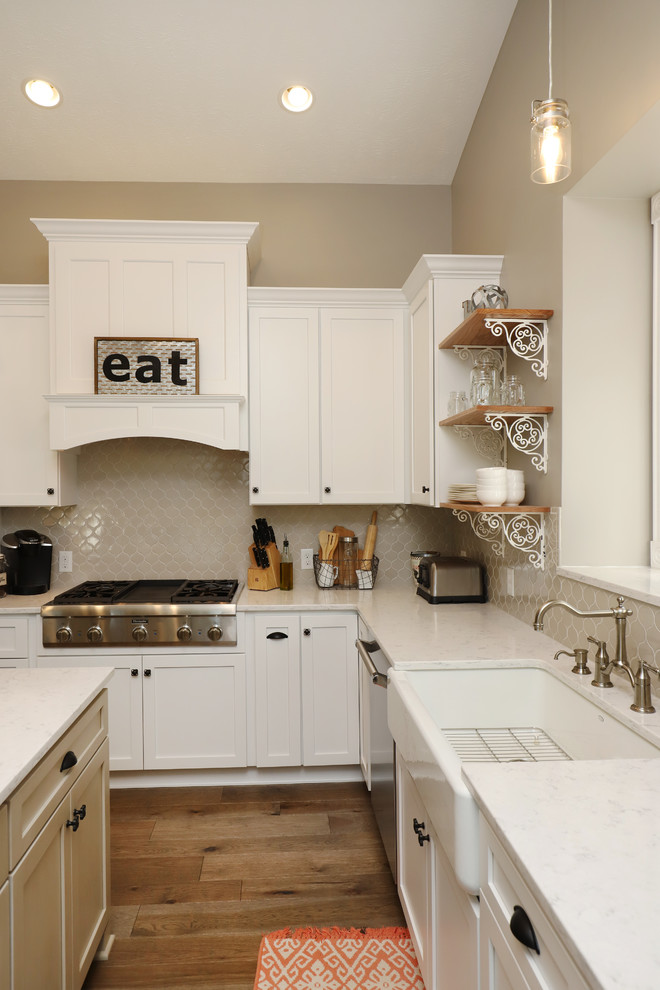 Design ideas for a medium sized farmhouse l-shaped kitchen/diner in Grand Rapids with a belfast sink, shaker cabinets, white cabinets, engineered stone countertops, grey splashback, ceramic splashback, stainless steel appliances, light hardwood flooring, an island and brown floors.
