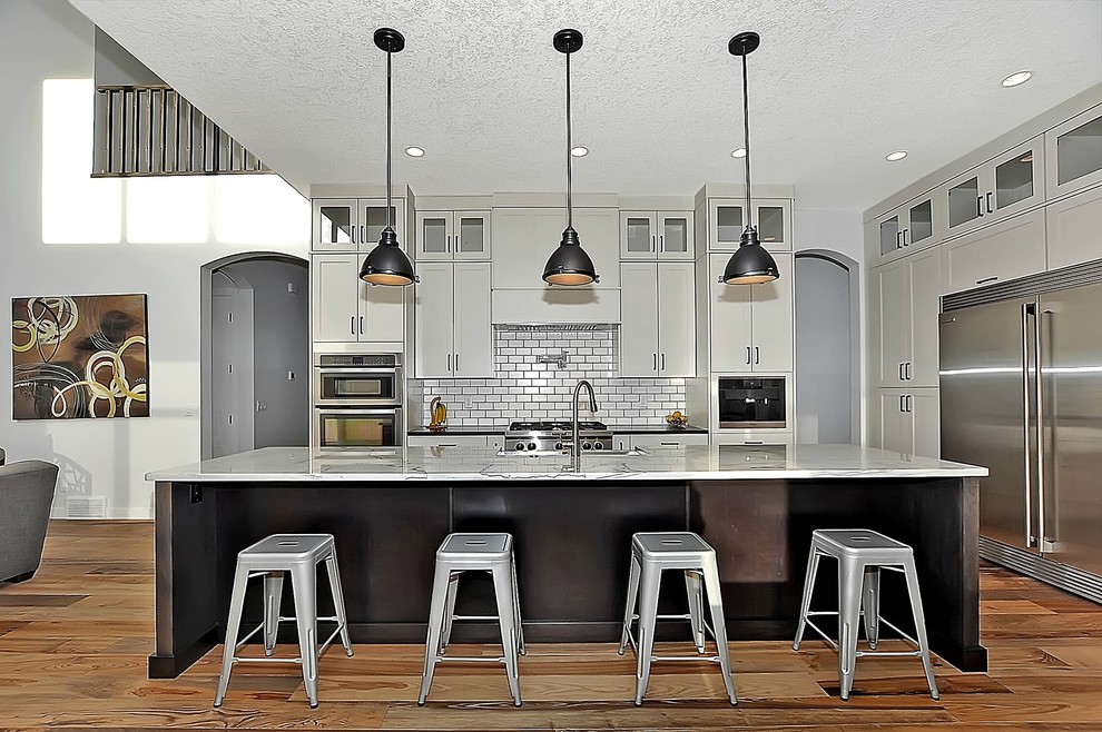 Example of a large country l-shaped medium tone wood floor eat-in kitchen design in Calgary with a farmhouse sink, shaker cabinets, granite countertops, white backsplash, ceramic backsplash, stainless steel appliances and an island