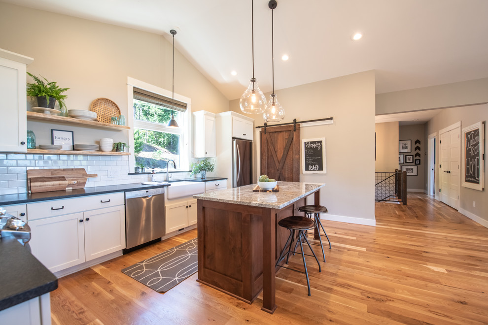 Mid-sized country l-shaped medium tone wood floor open concept kitchen photo in Seattle with a farmhouse sink, shaker cabinets, white cabinets, granite countertops, white backsplash, porcelain backsplash, stainless steel appliances, an island and black countertops