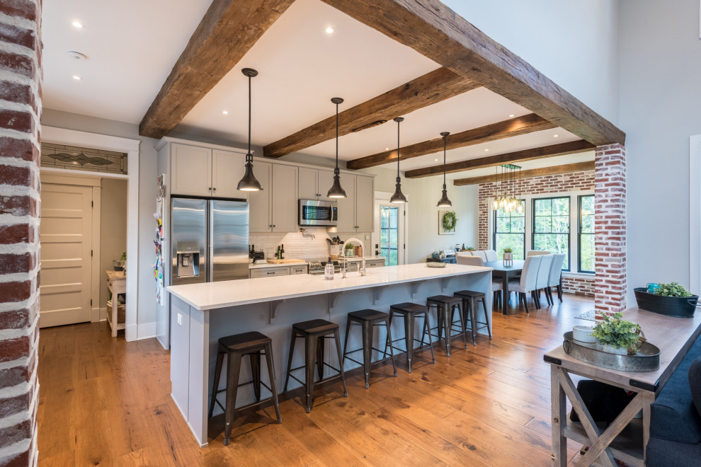 Example of a large cottage medium tone wood floor and brown floor eat-in kitchen design in Other with a farmhouse sink, recessed-panel cabinets, gray cabinets, quartz countertops, white backsplash, porcelain backsplash, stainless steel appliances, an island and white countertops