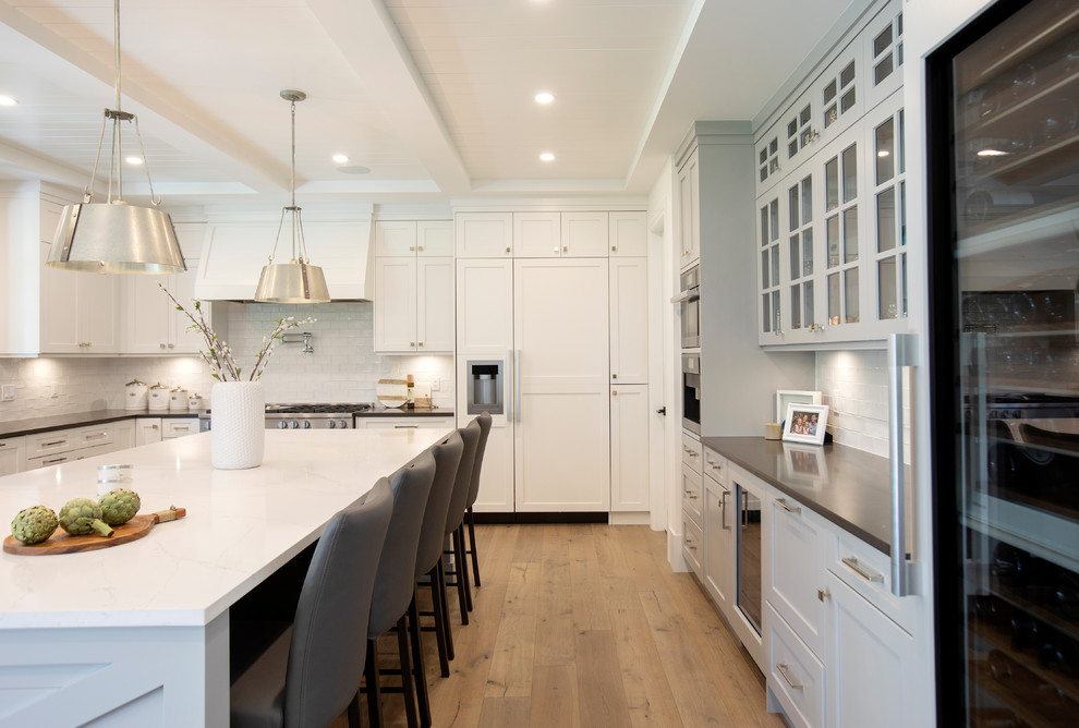 This is an example of a large classic u-shaped enclosed kitchen in Vancouver with white splashback, light hardwood flooring, an island, a belfast sink, shaker cabinets, white cabinets, metro tiled splashback, stainless steel appliances, brown floors and black worktops.