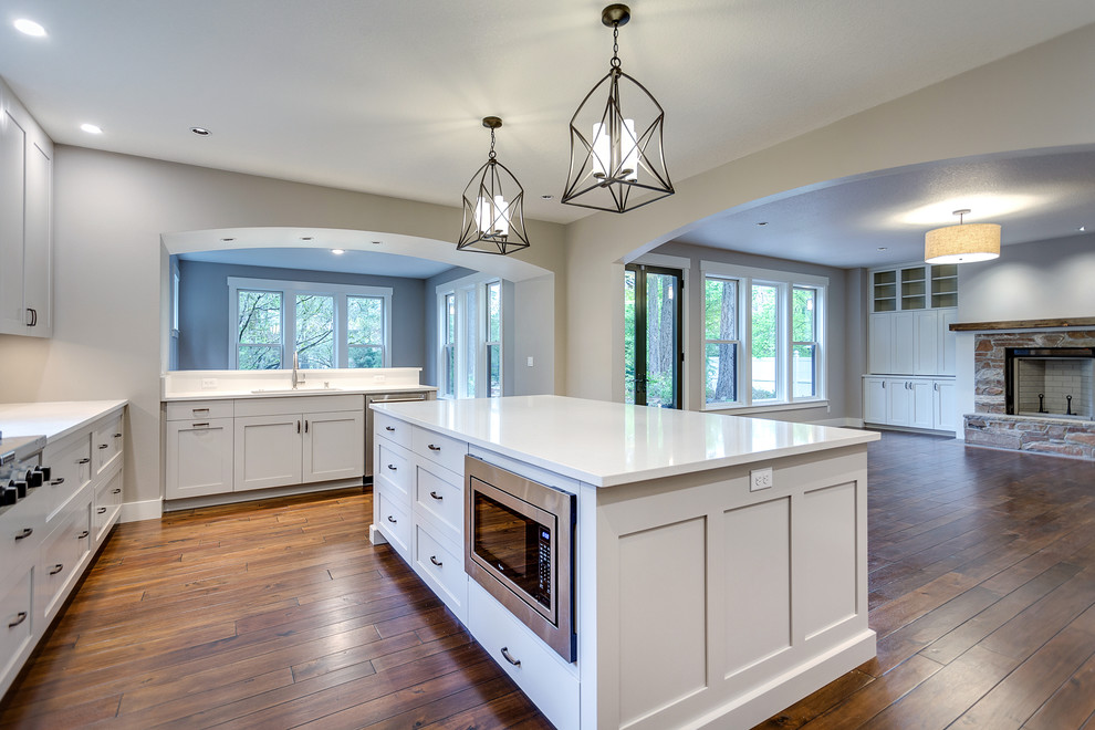 Bild på ett stort lantligt vit vitt kök, med en undermonterad diskho, luckor med infälld panel, grå skåp, bänkskiva i kvarts, grått stänkskydd, stänkskydd i stenkakel, rostfria vitvaror, mörkt trägolv, en köksö och brunt golv