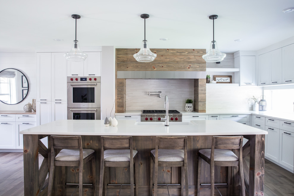 Inspiration for a country l-shaped dark wood floor and brown floor kitchen remodel in Other with a farmhouse sink, shaker cabinets, white cabinets, beige backsplash, stainless steel appliances, an island and white countertops