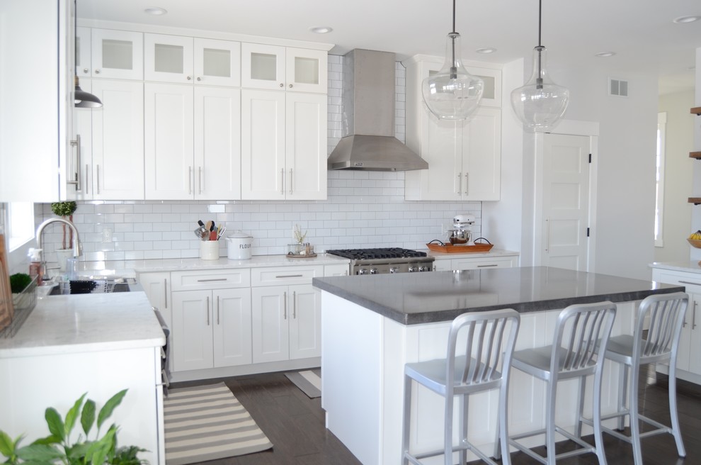 Photo of a medium sized rural l-shaped kitchen in Other with white cabinets, metro tiled splashback, an island, brown floors, a belfast sink, shaker cabinets, white splashback, stainless steel appliances and medium hardwood flooring.