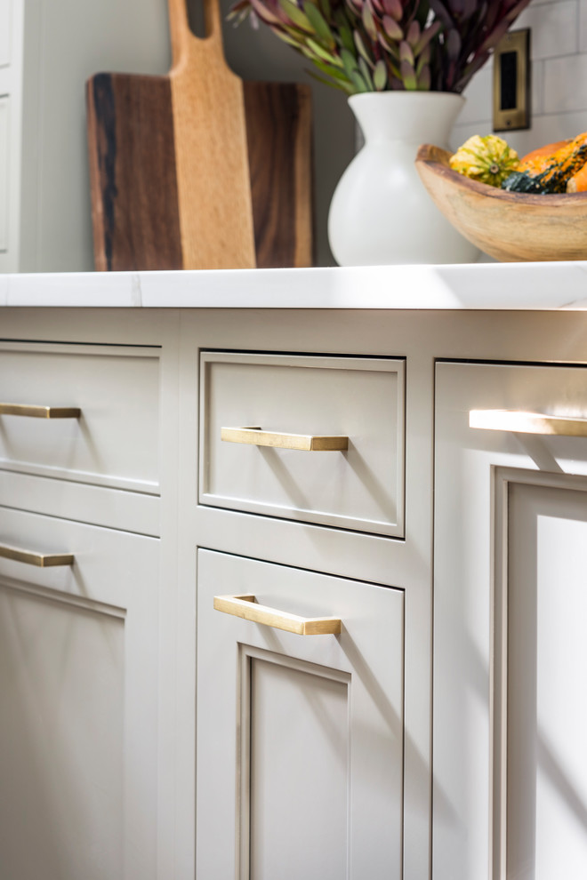 This is an example of a large country l-shaped kitchen/diner in New York with a double-bowl sink, shaker cabinets, beige cabinets, engineered stone countertops, white splashback, ceramic splashback, black appliances, medium hardwood flooring, an island, brown floors and white worktops.