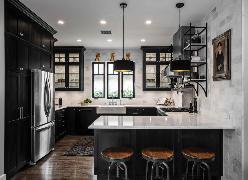 Medium sized farmhouse u-shaped enclosed kitchen in Los Angeles with a belfast sink, stainless steel appliances, a breakfast bar, brown floors, grey worktops, glass-front cabinets, black cabinets, grey splashback, metro tiled splashback, light hardwood flooring and engineered stone countertops.