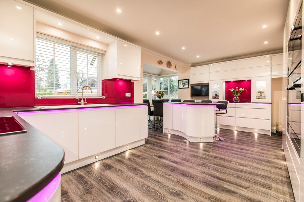 This is an example of a large modern u-shaped kitchen in Hampshire with an integrated sink, flat-panel cabinets, white cabinets, pink splashback, glass sheet splashback, stainless steel appliances, an island, brown floors and black worktops.