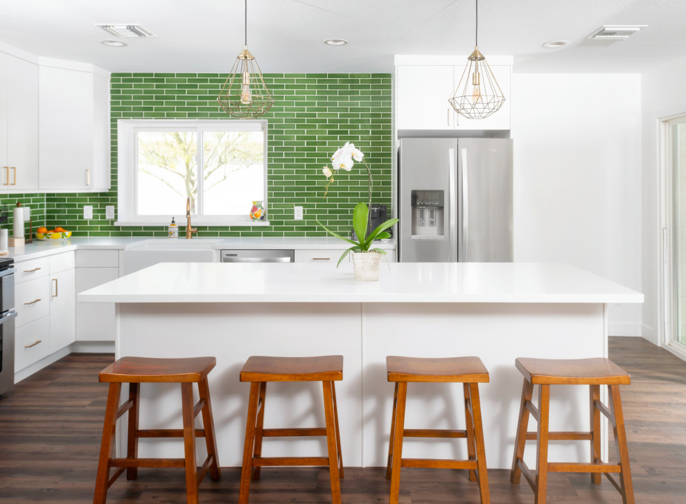Photo of a medium sized midcentury l-shaped open plan kitchen in Phoenix with a belfast sink, flat-panel cabinets, white cabinets, engineered stone countertops, green splashback, ceramic splashback, stainless steel appliances, vinyl flooring, an island, brown floors and white worktops.