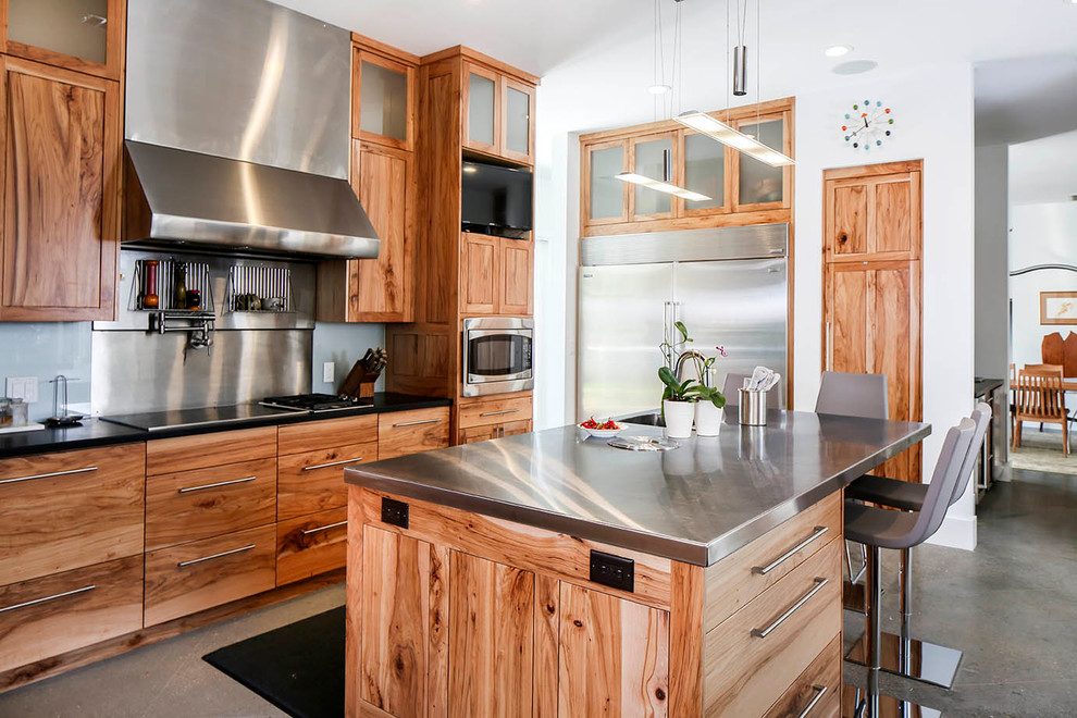 Modelo de cocina contemporánea con fregadero integrado, armarios con paneles lisos, puertas de armario de madera oscura, encimera de acero inoxidable, salpicadero metalizado, electrodomésticos de acero inoxidable, suelo de cemento y una isla