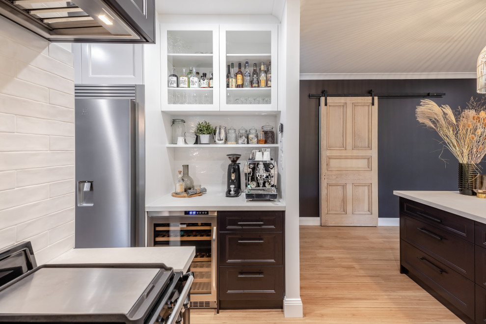 This is an example of a large traditional u-shaped kitchen pantry in Adelaide with a belfast sink, shaker cabinets, black cabinets, engineered stone countertops, white splashback, metro tiled splashback, black appliances, laminate floors, a breakfast bar, brown floors and grey worktops.