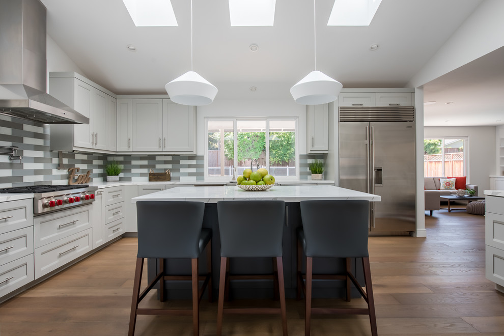 Mid-sized minimalist l-shaped medium tone wood floor and brown floor eat-in kitchen photo in San Francisco with a farmhouse sink, shaker cabinets, gray cabinets, granite countertops, gray backsplash, glass tile backsplash, stainless steel appliances, an island and white countertops