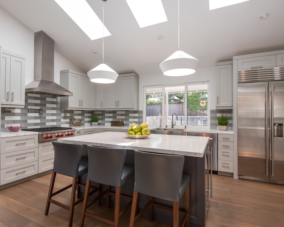 This is an example of a medium sized traditional l-shaped kitchen in San Francisco with a belfast sink, shaker cabinets, grey cabinets, granite worktops, grey splashback, glass tiled splashback, stainless steel appliances, medium hardwood flooring, an island, brown floors and white worktops.
