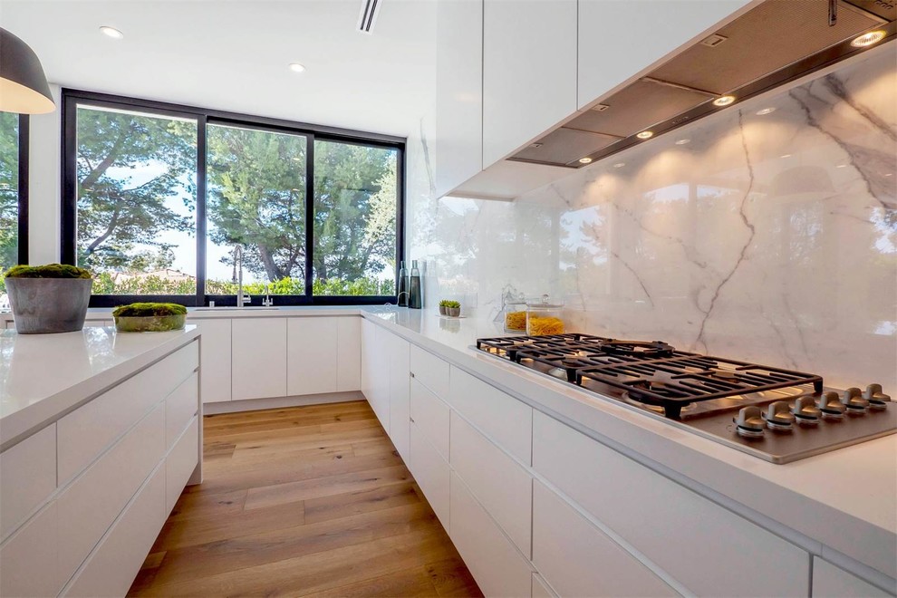 Large modern l-shaped open plan kitchen in Los Angeles with a submerged sink, flat-panel cabinets, white cabinets, composite countertops, multi-coloured splashback, marble splashback, stainless steel appliances, medium hardwood flooring, an island and brown floors.