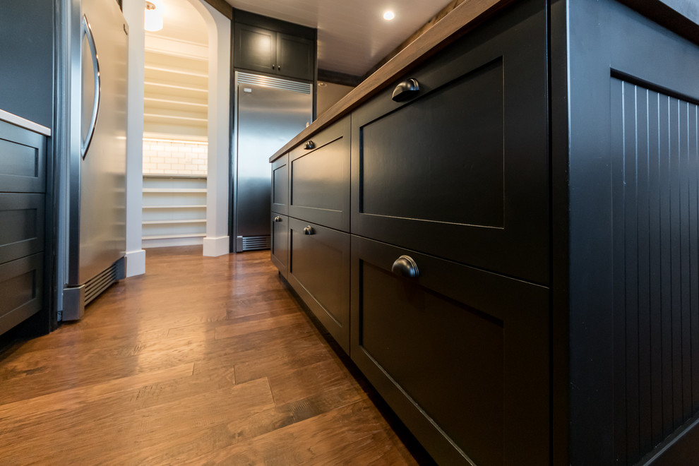 Inspiration for a large industrial u-shaped medium tone wood floor kitchen pantry remodel in Chicago with a farmhouse sink, shaker cabinets, black cabinets, wood countertops, white backsplash, subway tile backsplash, stainless steel appliances and an island