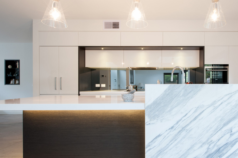 Large modern single-wall open plan kitchen in Melbourne with a submerged sink, flat-panel cabinets, white cabinets, engineered stone countertops, metallic splashback, mirror splashback, black appliances, porcelain flooring, an island and grey floors.