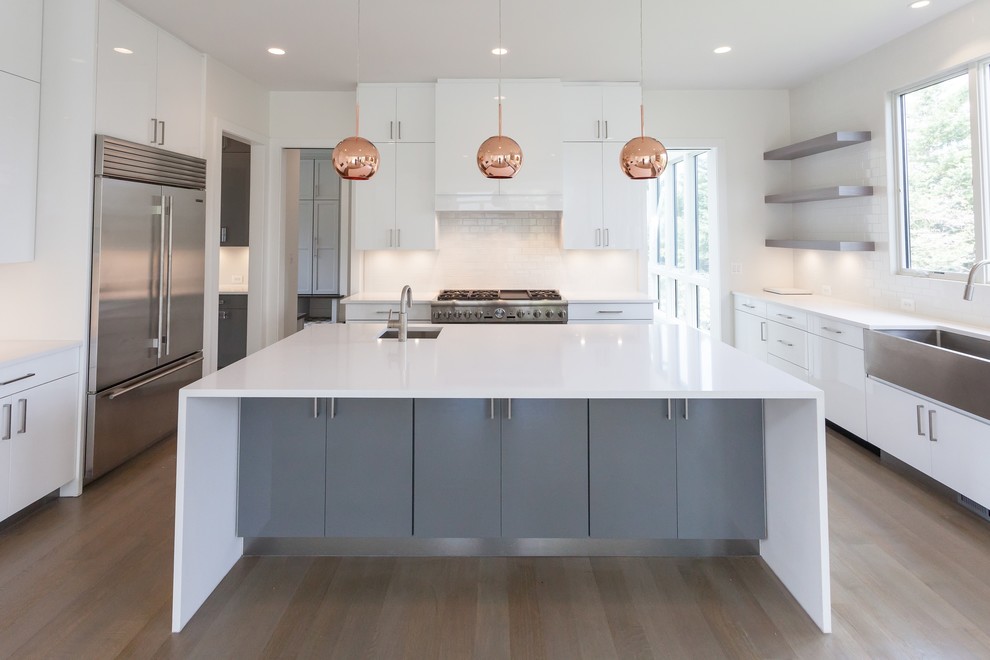 Contemporary u-shaped open plan kitchen in DC Metro with a belfast sink, flat-panel cabinets, white cabinets, white splashback, metro tiled splashback, stainless steel appliances, light hardwood flooring, an island, grey floors and white worktops.