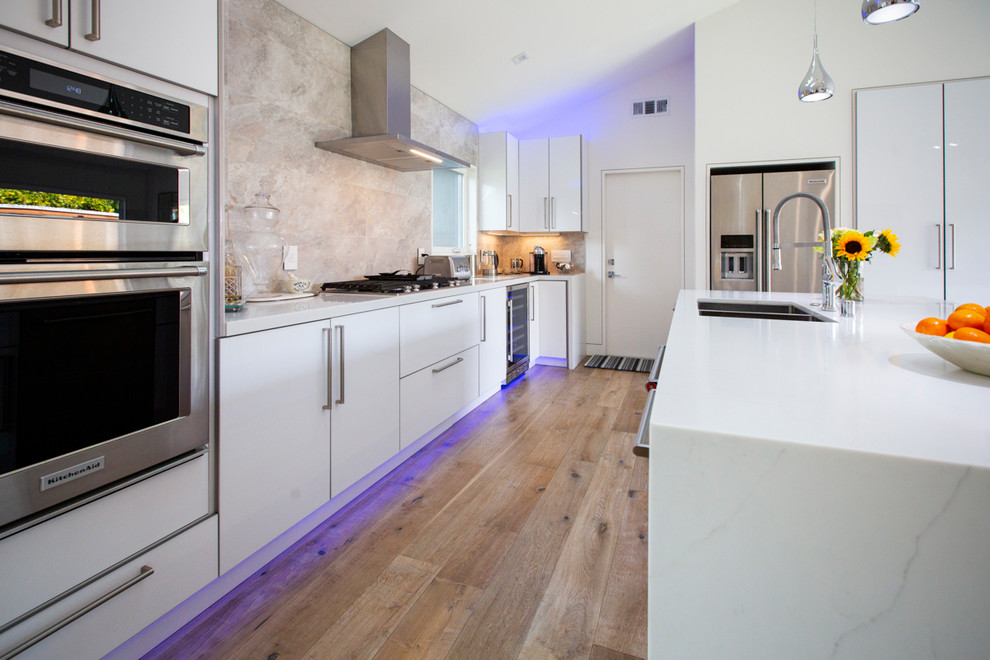 This is an example of a medium sized modern u-shaped open plan kitchen in San Francisco with a double-bowl sink, flat-panel cabinets, white cabinets, beige splashback, stainless steel appliances, light hardwood flooring, an island, brown floors, white worktops, quartz worktops and limestone splashback.