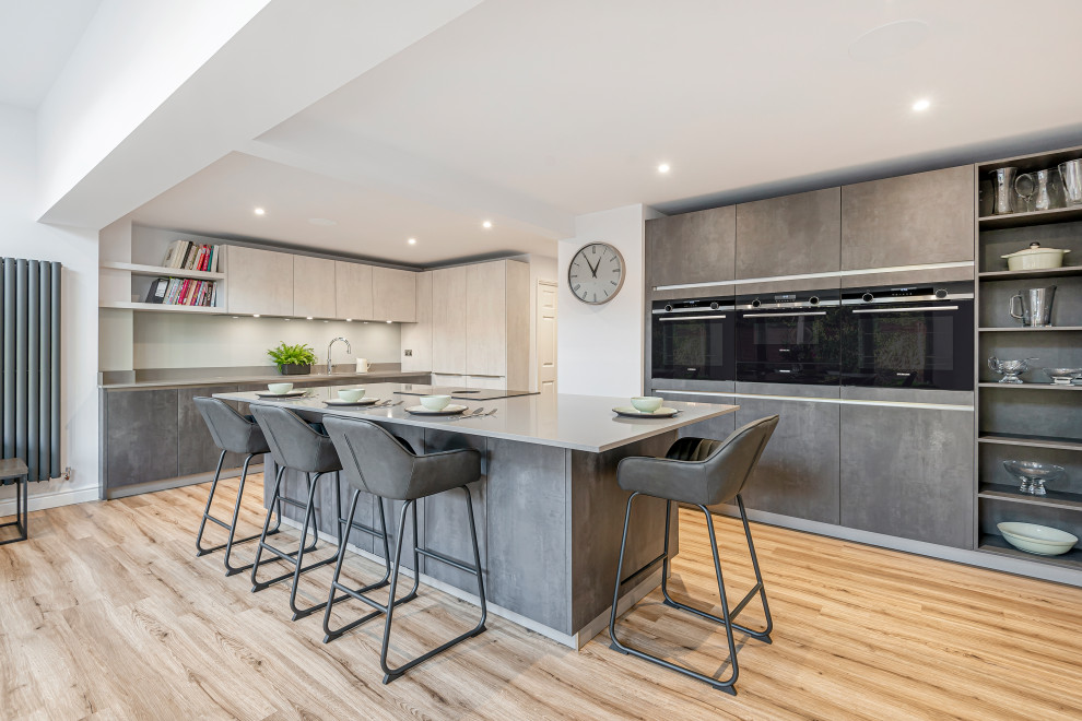 Inspiration for a large contemporary l-shaped open plan kitchen in Berkshire with flat-panel cabinets, an island, grey cabinets, composite countertops, grey splashback, black appliances, light hardwood flooring, beige floors and grey worktops.