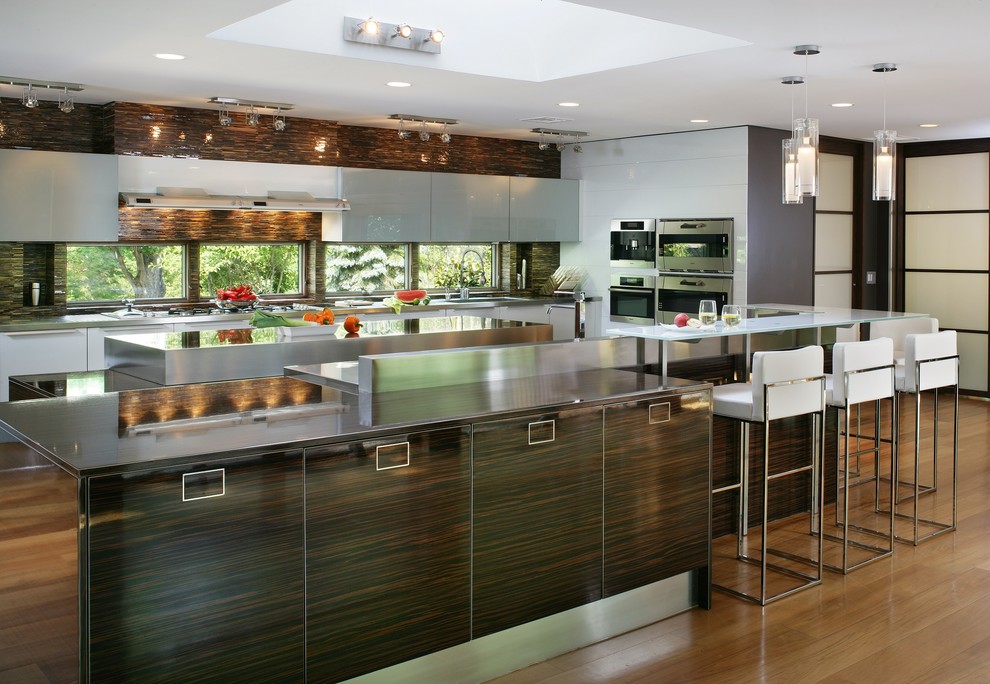 Inredning av ett modernt stort kök, med en integrerad diskho, släta luckor, grå skåp, bänkskiva i rostfritt stål, brunt stänkskydd, stänkskydd i glaskakel, rostfria vitvaror, mellanmörkt trägolv och flera köksöar
