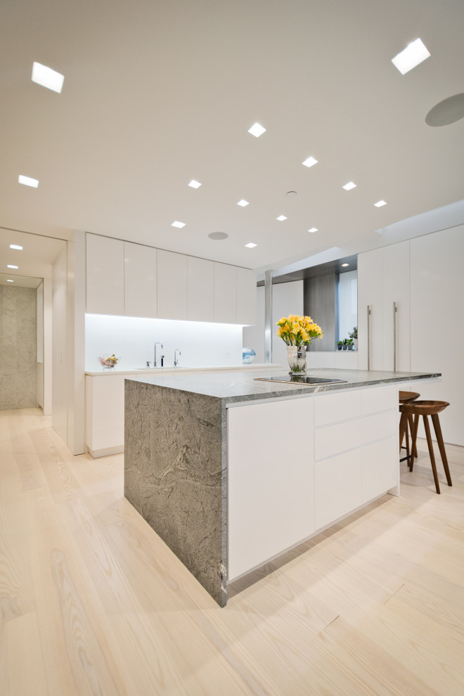 This is an example of a modern l-shaped kitchen in New York with a submerged sink, flat-panel cabinets, white cabinets, marble worktops, white splashback, stone slab splashback, light hardwood flooring, an island, green worktops, integrated appliances and beige floors.