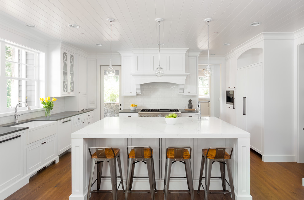 Large country u-shaped kitchen/diner in DC Metro with a belfast sink, recessed-panel cabinets, white cabinets, engineered stone countertops, white splashback, metro tiled splashback, integrated appliances, dark hardwood flooring, an island, brown floors and grey worktops.