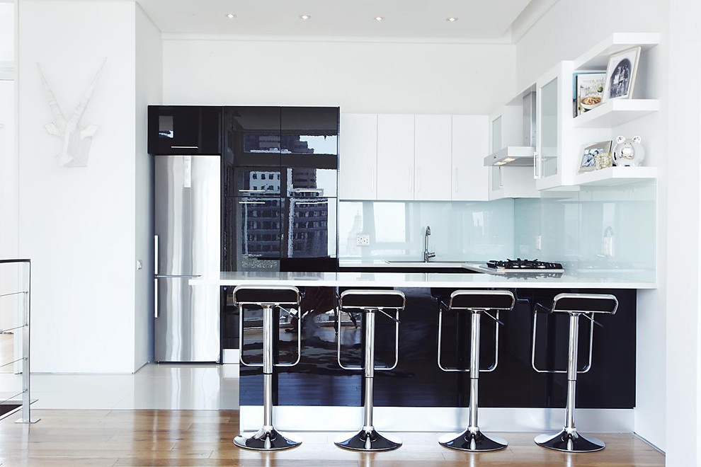 На фото: п-образная, глянцевая кухня в стиле модернизм с плоскими фасадами, черными фасадами, фартуком из стекла и техникой из нержавеющей стали