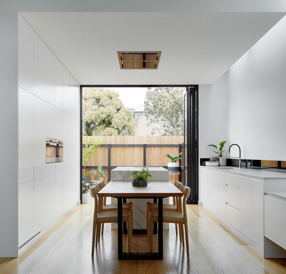 На фото: параллельная кухня в стиле модернизм с обеденным столом, врезной мойкой, плоскими фасадами, белыми фасадами, светлым паркетным полом, островом и бежевым полом с