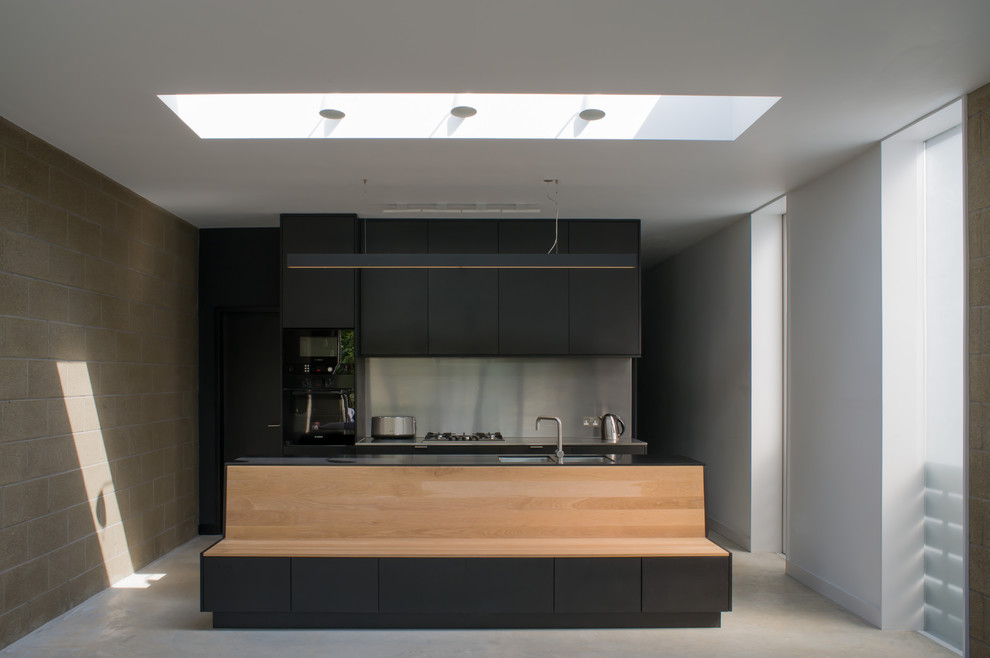 Design ideas for a modern galley kitchen/diner in London with a double-bowl sink, flat-panel cabinets, black cabinets, composite countertops, metallic splashback, black appliances, concrete flooring and an island.