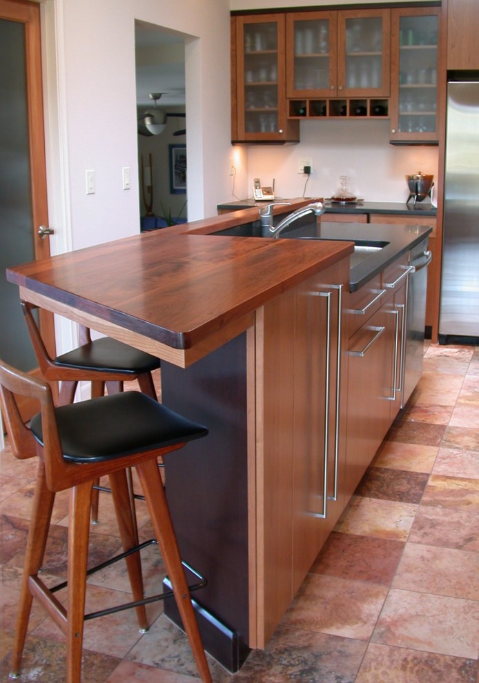 Cette image montre une cuisine américaine minimaliste en L et bois brun avec un plan de travail en bois, un évier encastré, une crédence en carrelage de pierre et un électroménager en acier inoxydable.
