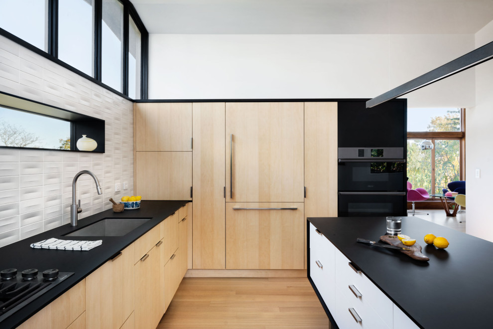 Photo of a contemporary kitchen in New York with a submerged sink, flat-panel cabinets, light wood cabinets, laminate countertops, white splashback, ceramic splashback, black appliances, light hardwood flooring, an island, black worktops and a vaulted ceiling.