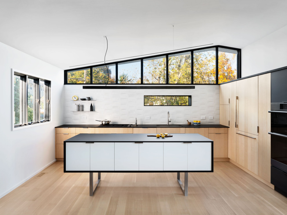 Inspiration for a contemporary kitchen in New York with a submerged sink, flat-panel cabinets, light wood cabinets, laminate countertops, white splashback, ceramic splashback, black appliances, light hardwood flooring, an island, black worktops and a vaulted ceiling.