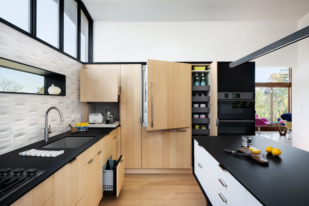 Photo of a modern kitchen in New York with a submerged sink, flat-panel cabinets, light wood cabinets, laminate countertops, white splashback, ceramic splashback, black appliances, light hardwood flooring, an island, black worktops and a vaulted ceiling.