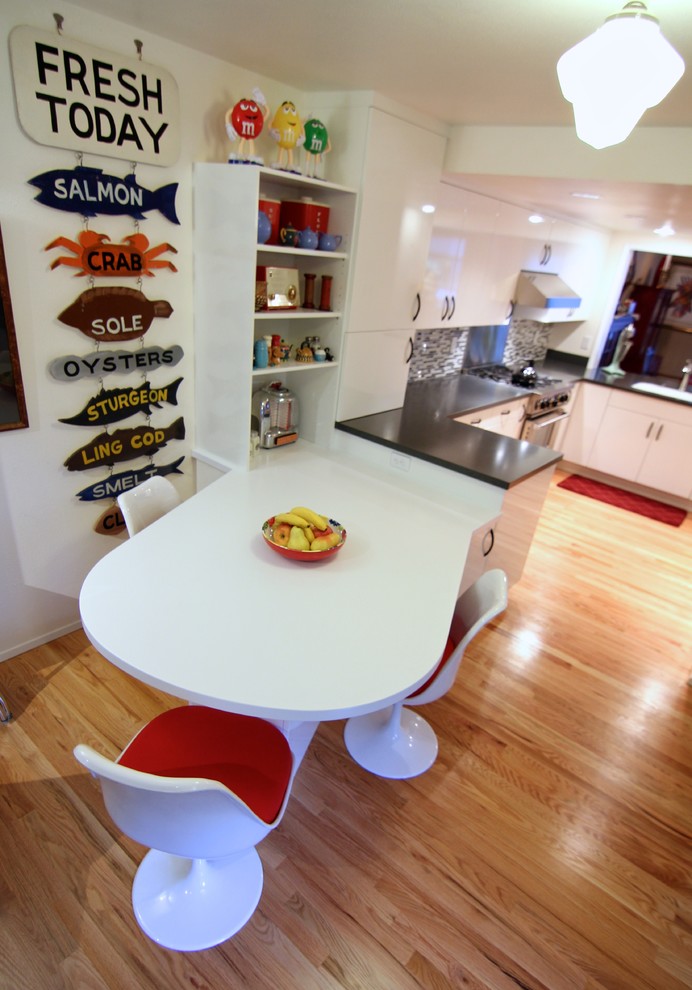 Medium sized eclectic galley kitchen/diner in Seattle with a built-in sink, flat-panel cabinets, white cabinets, laminate countertops, grey splashback, glass tiled splashback, stainless steel appliances, light hardwood flooring and a breakfast bar.