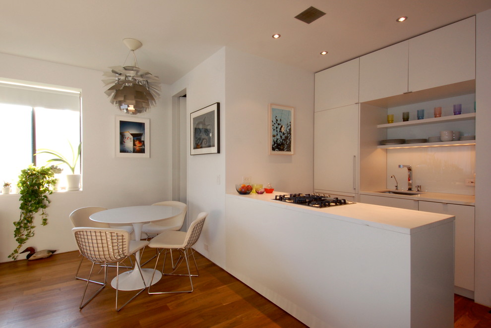 Стильный дизайн: параллельная кухня в стиле модернизм с обеденным столом, одинарной мойкой, плоскими фасадами, белыми фасадами, белым фартуком, фартуком из стекла и техникой под мебельный фасад - последний тренд