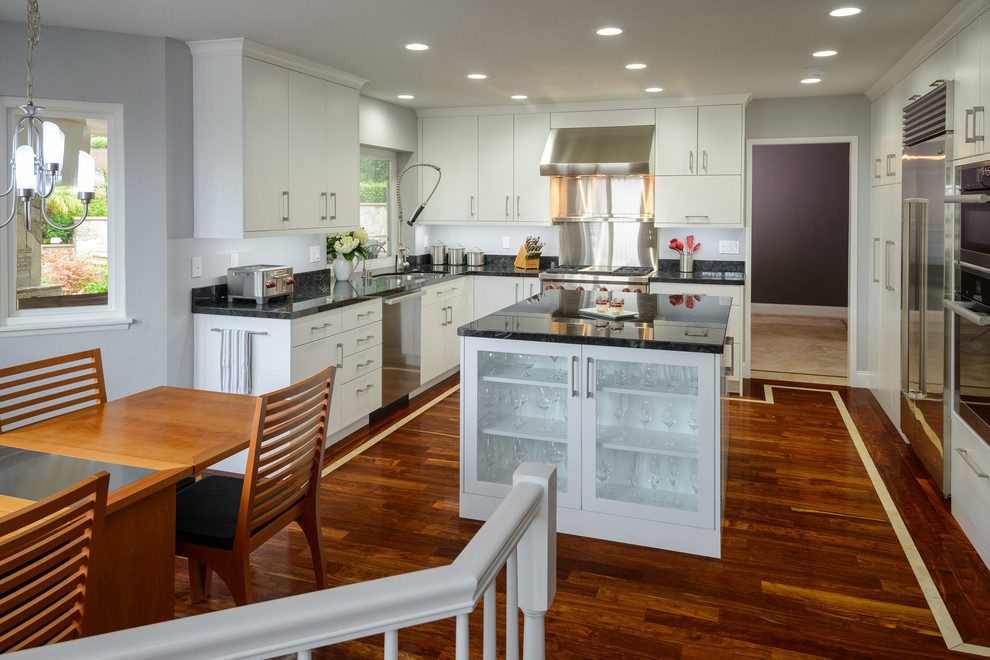 Bild på ett stort funkis kök, med en nedsänkt diskho, släta luckor, vita skåp, rostfria vitvaror, mellanmörkt trägolv, en köksö, granitbänkskiva, svart stänkskydd och stänkskydd i skiffer